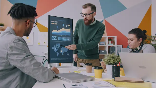 A man pointing at a vertical monitor displaying data charts to two colleagues Young man is making presentation using digital screen talking to group of colleagues in office. Modern. Analytics tools: what beginners should know